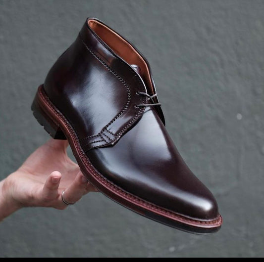 A hand showcases the Alden Madison D5706C - Antique Chukka Boot in Color 8 Shell Cordovan, featuring polished dark brown leather, black laces, and detailed stitching against a gray background.