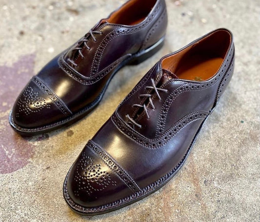The Alden Madison 9854 Medallion Cap Toe Bal in Color 8 Shell Cordovan features a polished finish, brogue detailing, and a classic leather sole, displayed on a textured concrete surface.
