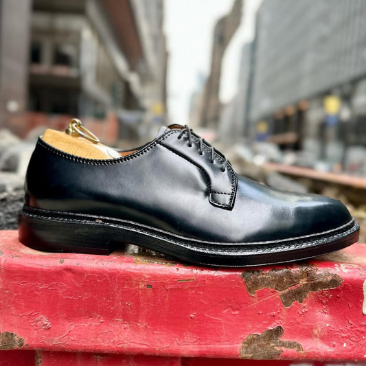 A single Alden Madison 9901 Plain Toe Blucher in black Shell Cordovan with a Double Oak Leather Sole sits on a worn red barricade, set against a blurred city street and towering buildings in the background.