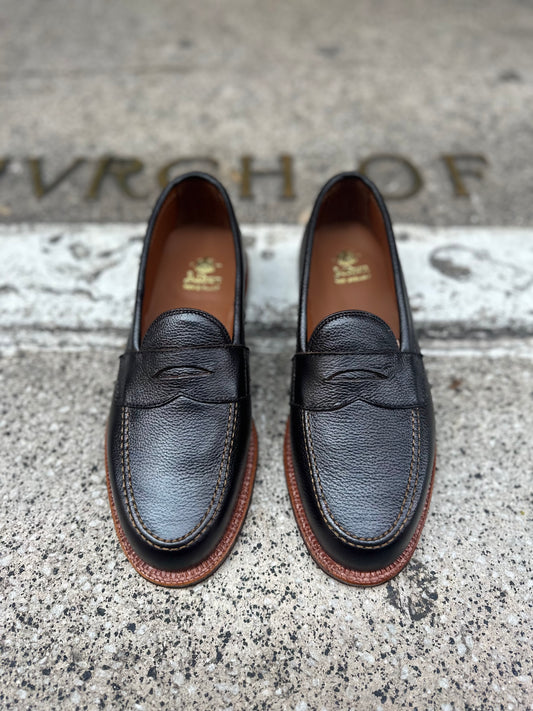 A pair of Alden Madisons 62456F - Penny Loafer in Brown Regina Grain sits on a textured concrete surface. The words VRCH OF are partially engraved behind them, adding an air of mystery to these black leather loafers with brown soles.