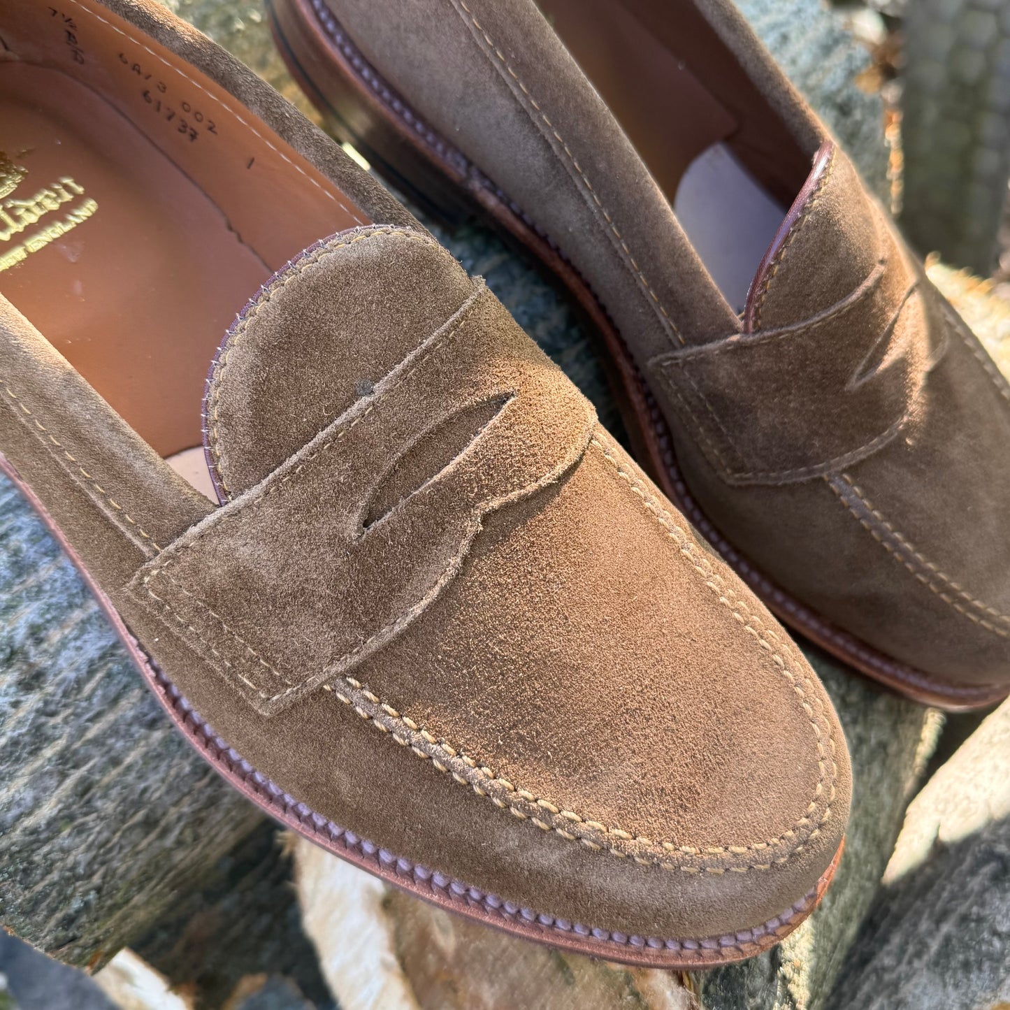 Close-up of Alden Madison 6173F - Penny Loafer in Snuff Suede, showing visible stitching on a stone surface. The shoes feature soft suede, leather lining, and a Flex Leather Sole for comfort.