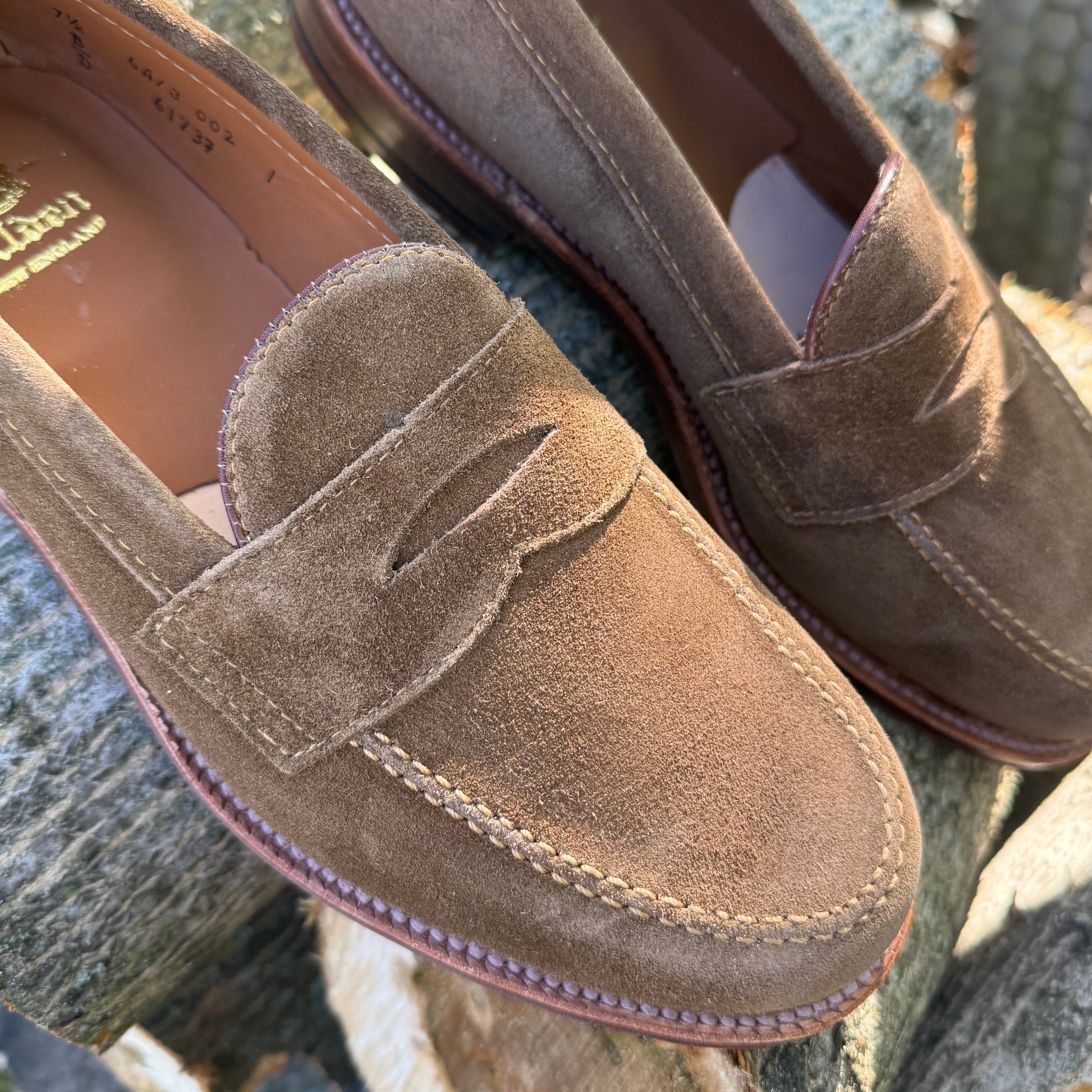 Close-up of Alden Madison 6173F - Penny Loafer in Snuff Suede, showing visible stitching on a stone surface. The shoes feature soft suede, leather lining, and a Flex Leather Sole for comfort.