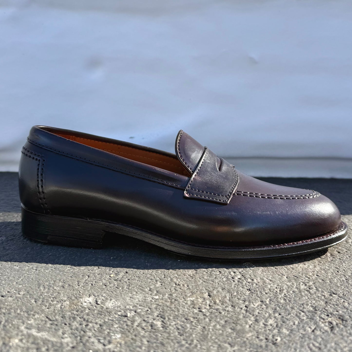 The ALDEN SHOES 96948 - Copley Penny Loafer in Color 8 Shell features dark brown leather, visible stitching, a penny strap, leather sole, and low heel; showcased on a textured gray surface with a blurred white background.