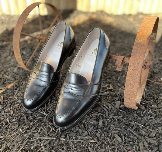 The Alden Madison 681 Full Strap Loafer in Black Calfskin, featuring leather soles, is showcased outdoors on dark mulch between two rusty metal wheels, with sunlight highlighting their polished shine.