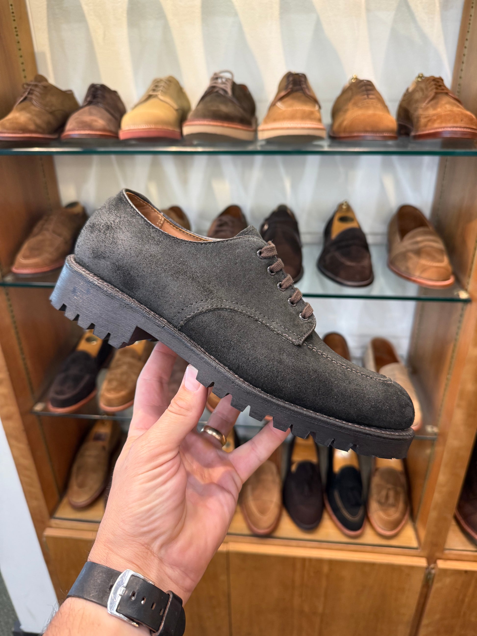 A hand holds the ALDEN SHOES 4006 - Earth Reverse Indy Shoe Heavy Lug, featuring black suede, a heavy lug rubber sole, and Trubalance Last, in a shoe store. Several pairs of brown and tan shoes are displayed on glass shelves in the background.