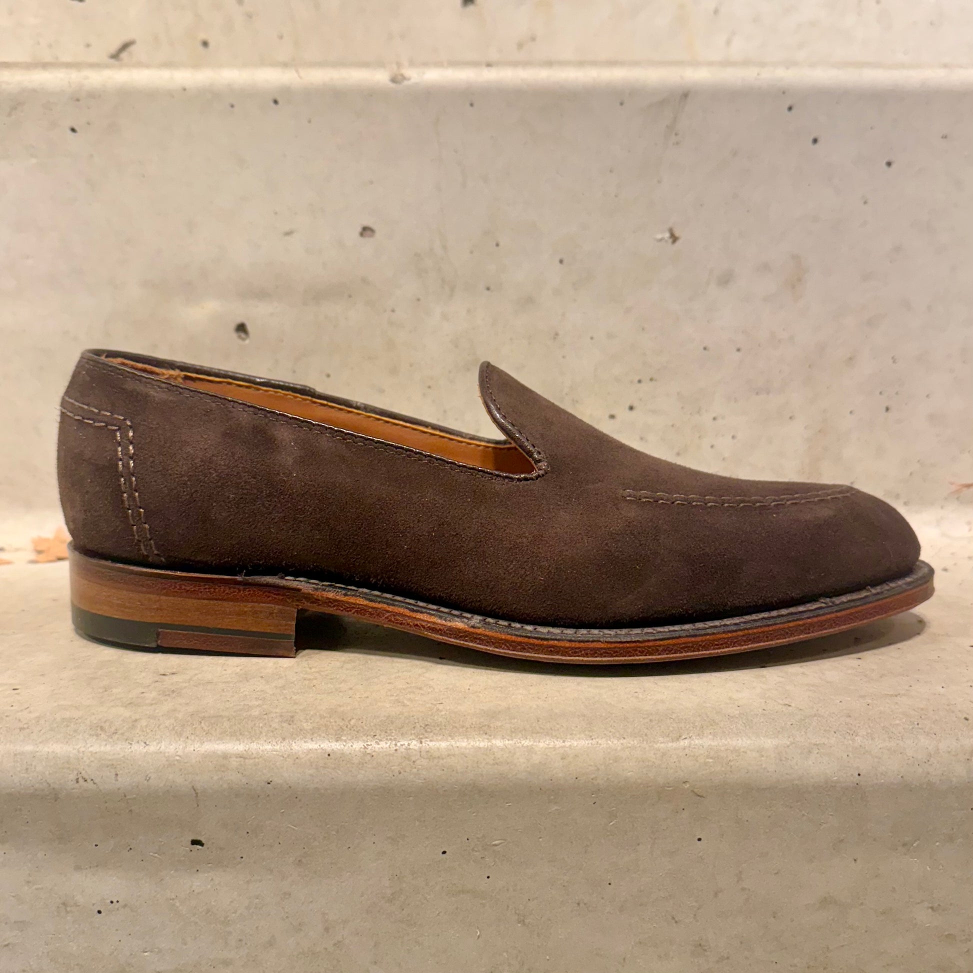 The ALDEN SHOES D5105 Vanderbilt in Dark Brown Suede, featuring white stitching and a flex leather sole, rests on a light-colored stone step.