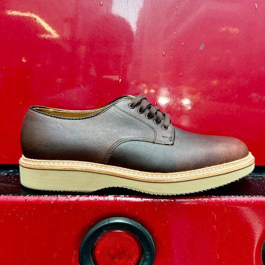 The Alden Madison 29401 “Ruslan” Plain Toe Dover in brown Kudu leather, featuring a tan wedge rubber sole, is shown side-on against a glossy red background with water droplets, highlighting its stitching and textured sole.