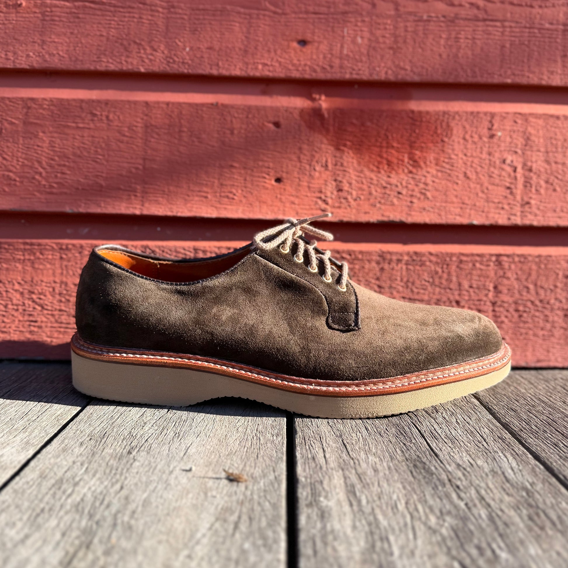 The ALDEN SHOES D5426 - Loden Suede Plain Toe Blucher, featuring a brown suede upper and tan wedge sole, is shown on wooden planks with a red wooden wall in bright sunlight.
