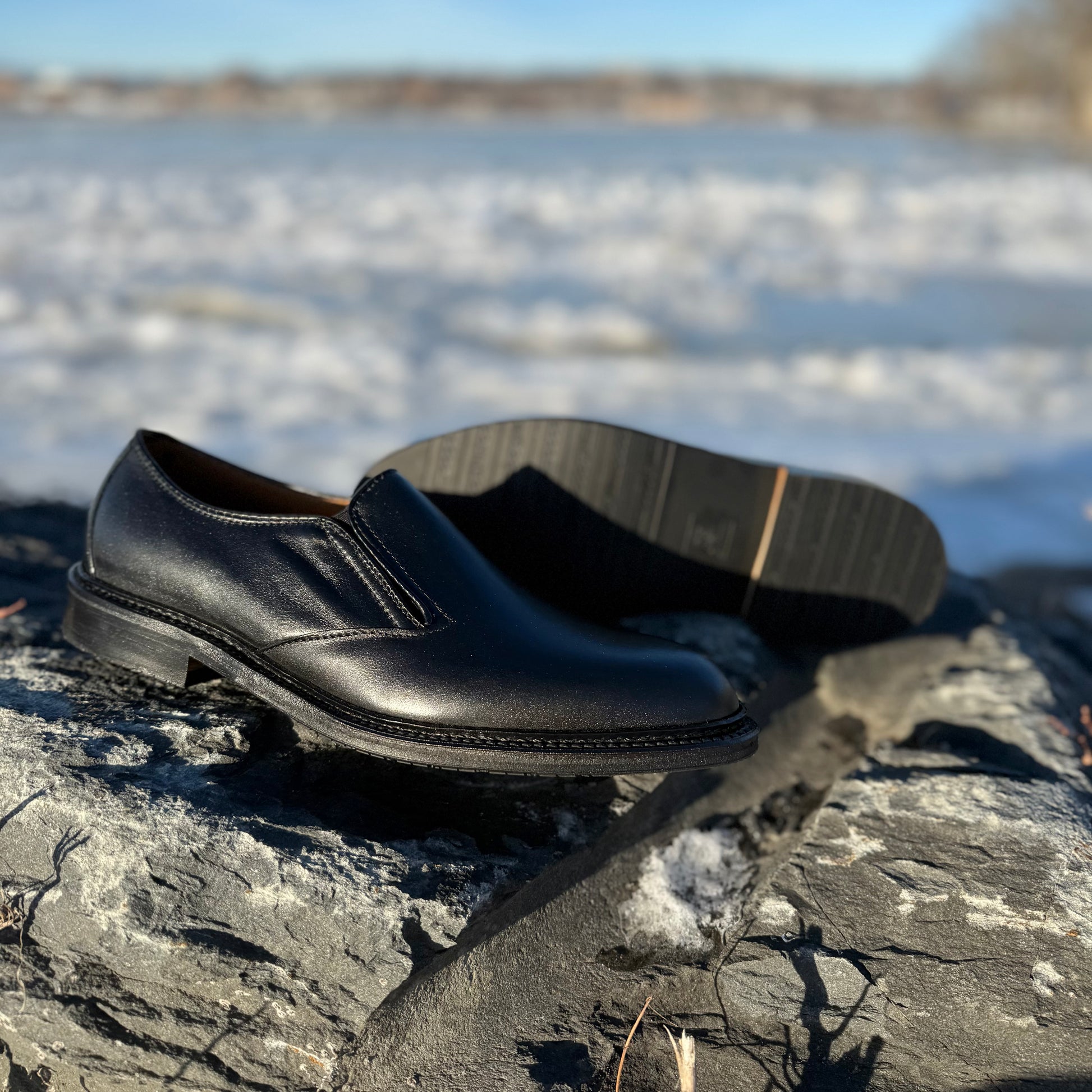 A pair of ALDEN SHOES D2406L "Double Gore" Slip On Rubber Sole dress shoes are displayed on a large gray rock outdoors, set against a blurred, snow-covered landscape and blue sky in the background.