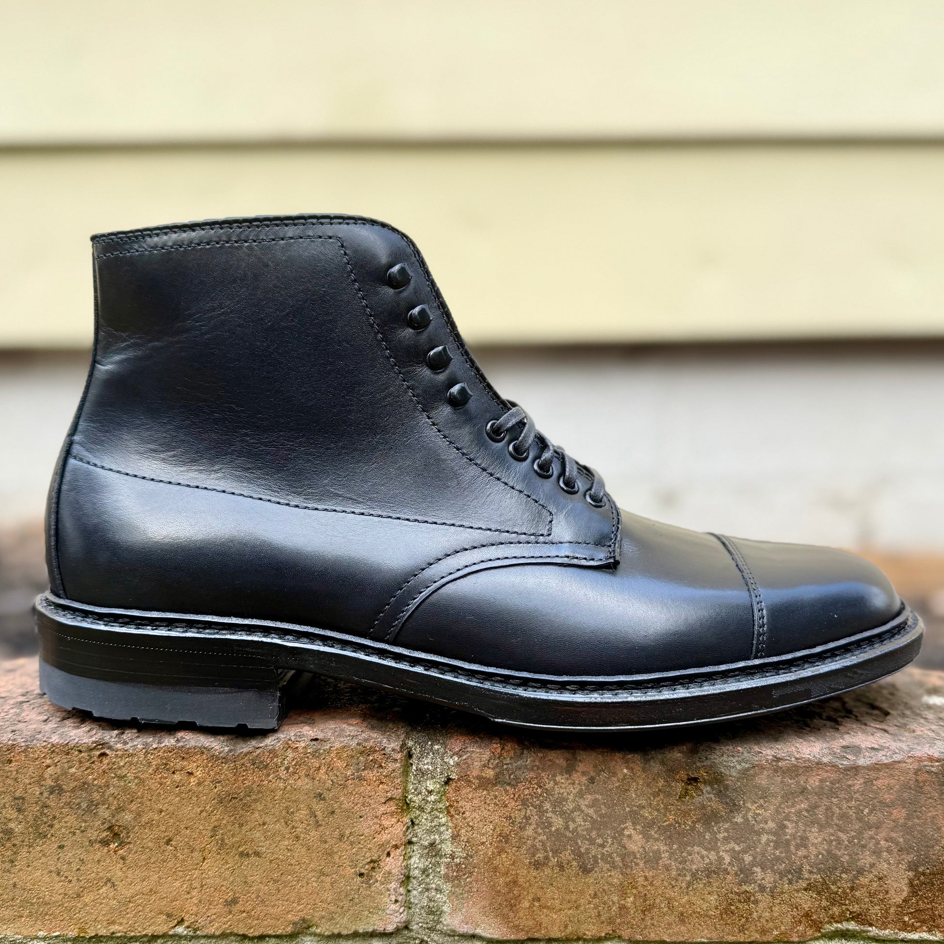 The Alden Madison 40797HC Black Cap Toe Boot with Commando Sole, crafted in calfskin with a lace-up design and Barrie last, is shown in profile on a brick ledge against a blurred beige background.