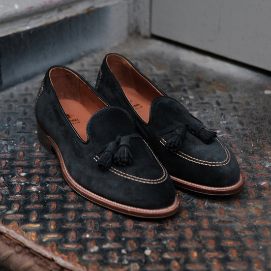 The Alden Madison D0106 Black Suede Tassel Loafers, featuring brown leather soles and white stitching, are displayed on a textured metal surface near a gray door.