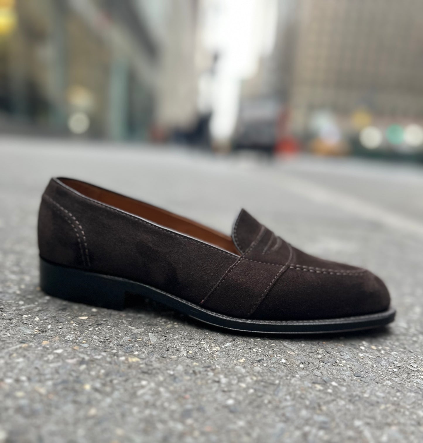 The ALDEN SHOES G9201 - Dark Brown Suede Full Strap Loafer, crafted on the Aberdeen last, rests on a city street with blurred buildings and traffic in the background.