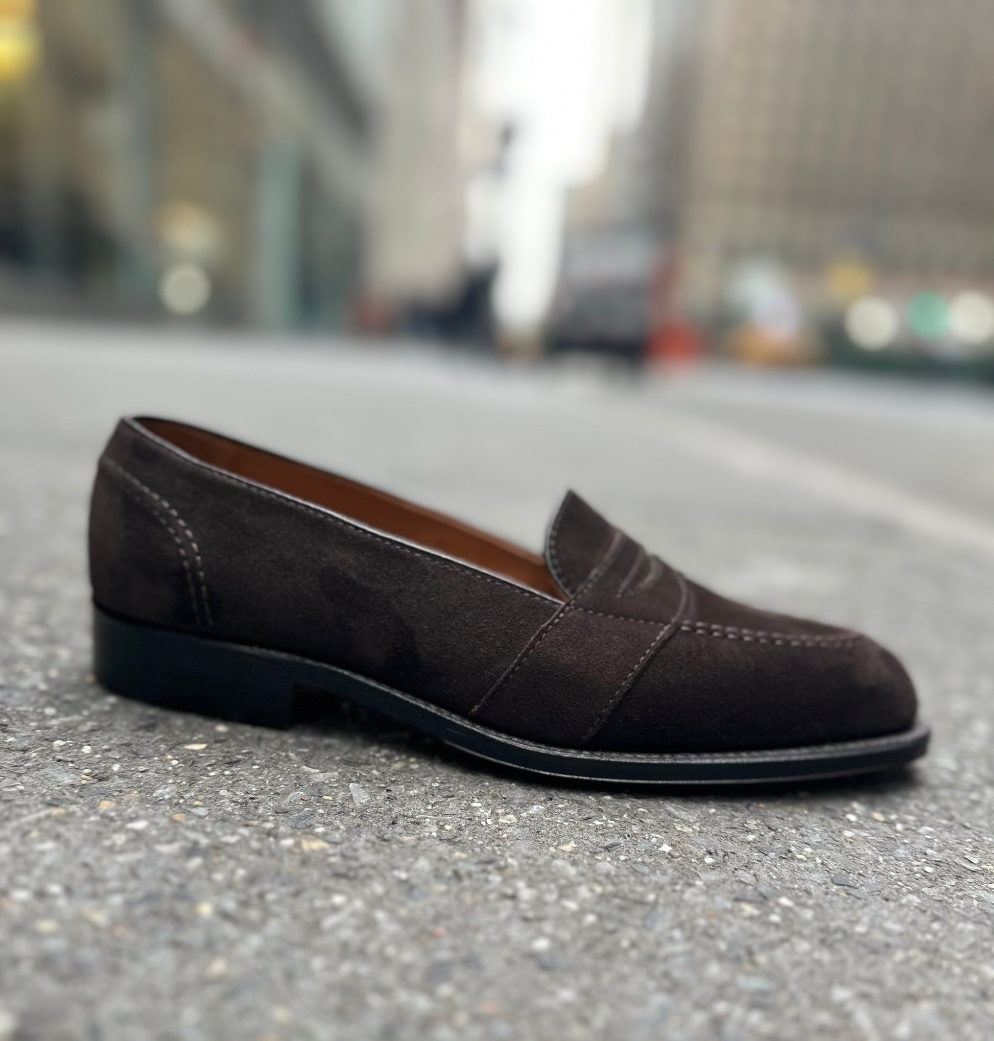 The ALDEN SHOES G9201 - Dark Brown Suede Full Strap Loafer, crafted on the Aberdeen last, rests on a city street with blurred buildings and traffic in the background.