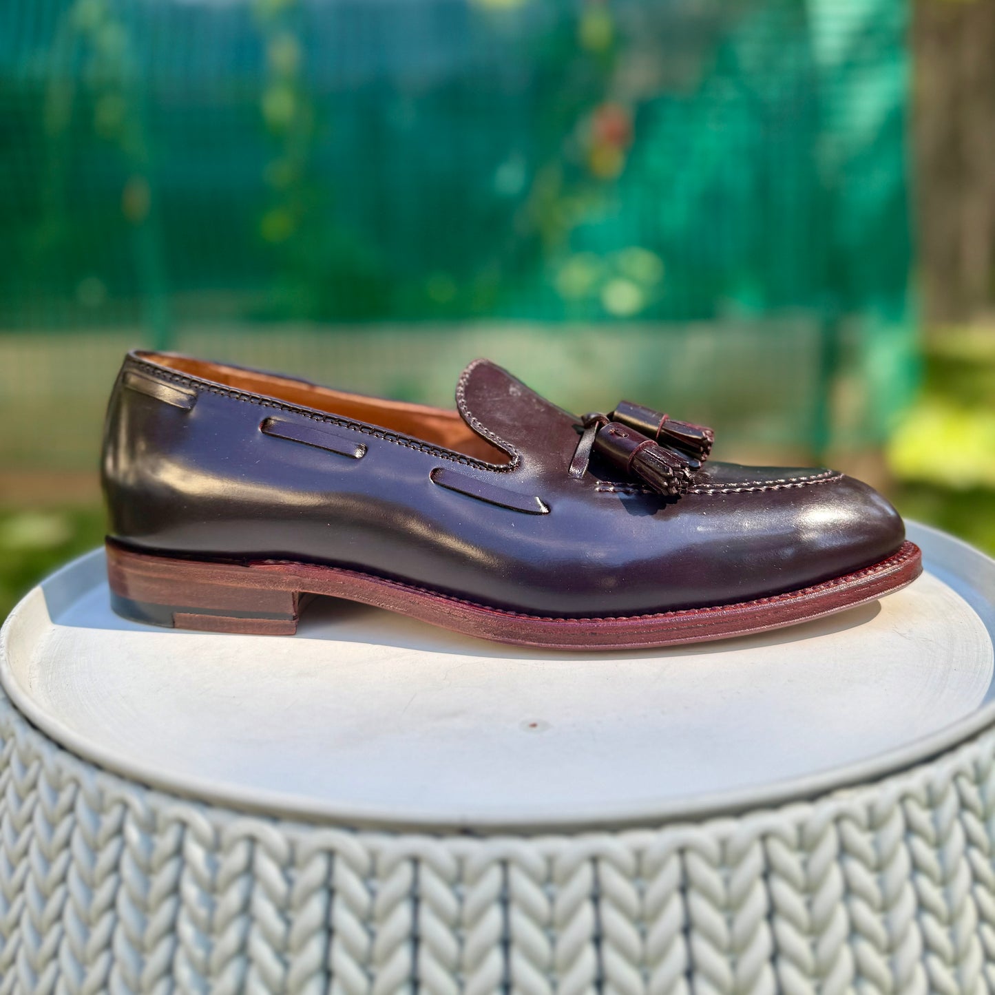 The Alden Madison D7103 - Antique Tassel Loafer in Color 8 Shell Cordovan rests on a round, woven white surface with a blurred green outdoor background.