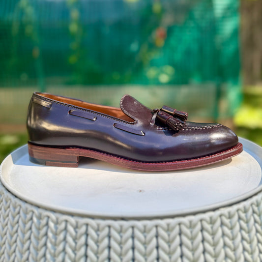 The Alden Madison D7103 - Antique Tassel Loafer in Color 8 Shell Cordovan rests on a round, woven white surface with a blurred green outdoor background.