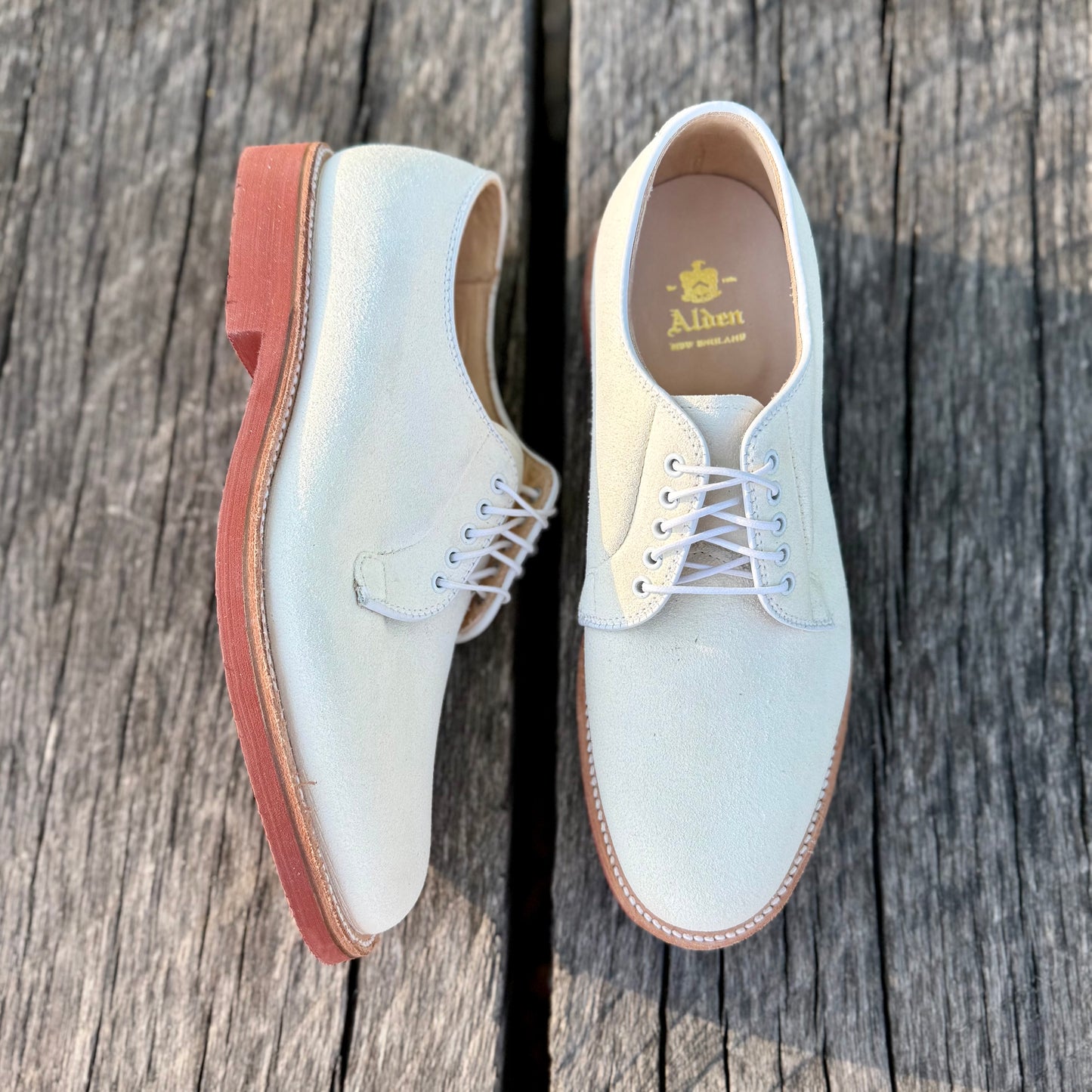 Alden Madison’s 9448 - White Buck in Ivory Suede lace-up dress shoes, built on the Barrie Last with reddish-brown soles, are shown on weathered wooden planks.