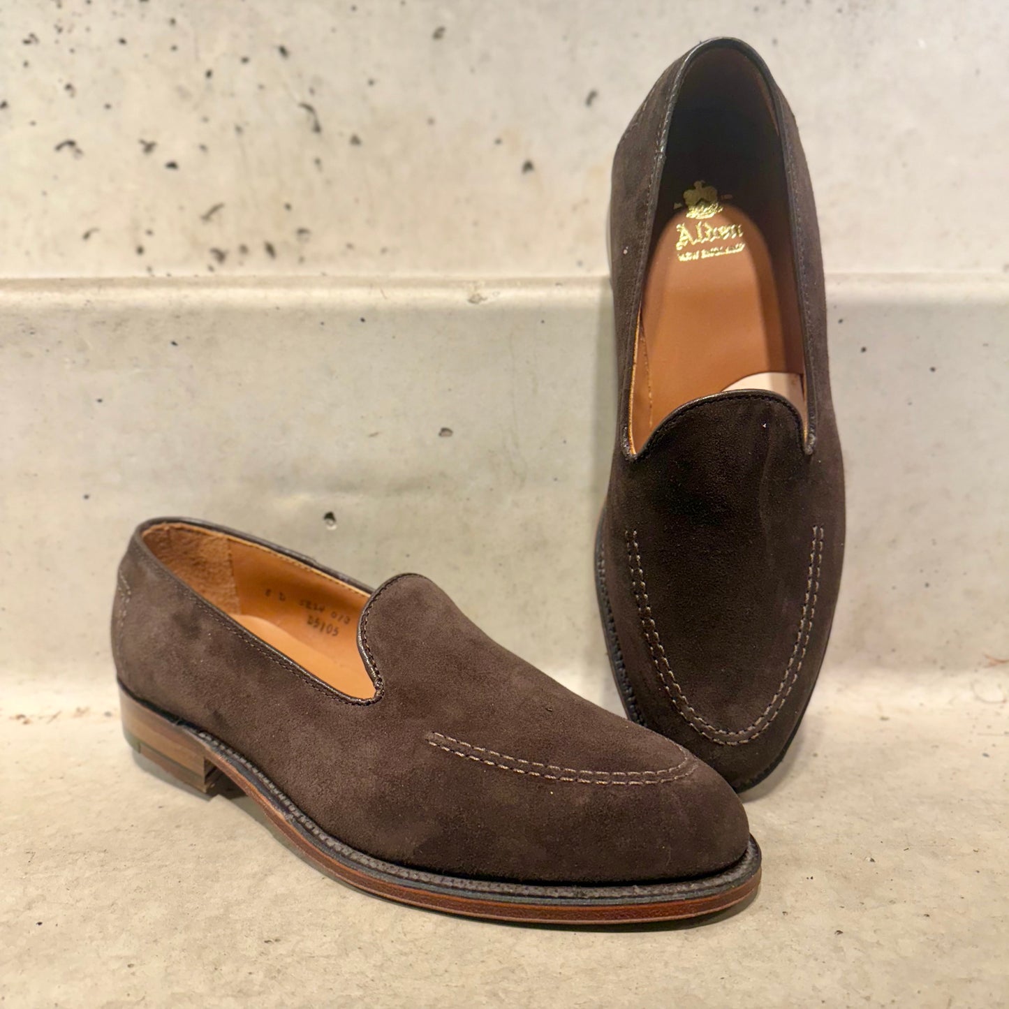 A pair of ALDEN SHOES D5105 - Vanderbilt in Dark Brown Suede with stitching detail, one upright and one on its side, featuring a Flex Leather Sole, displayed on a light textured surface.