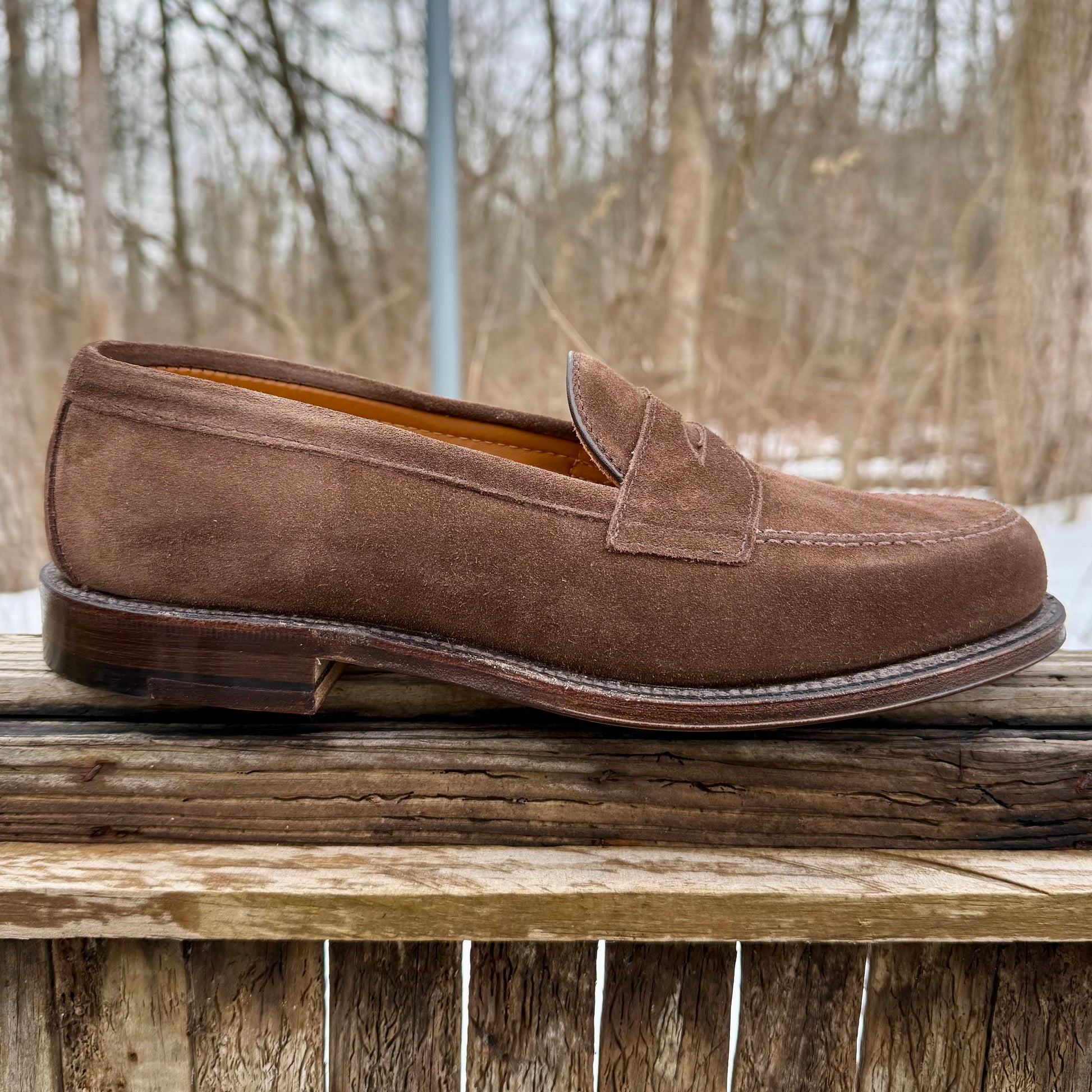 The Alden Madison 6175F Penny Loafer in Humus Suede, featuring a Flex Oiled Leather Sole and low heel, rests on a weathered wooden surface with leafless trees and an overcast sky in the background.