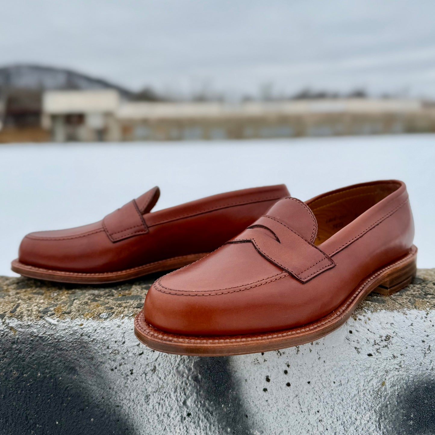 The Alden Madison 9823 - Penny Loafer in Tan Michigan Calf with an oak leather sole is displayed outdoors on concrete, showcasing its classic design against a blurred snowy backdrop with buildings.