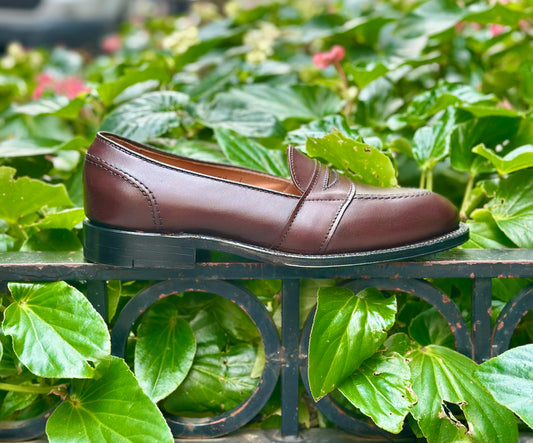 The Alden Madison 686 Full Strap Loafer in brown calfskin is shown on a metal railing, set against vibrant green leaves and plants in the background.