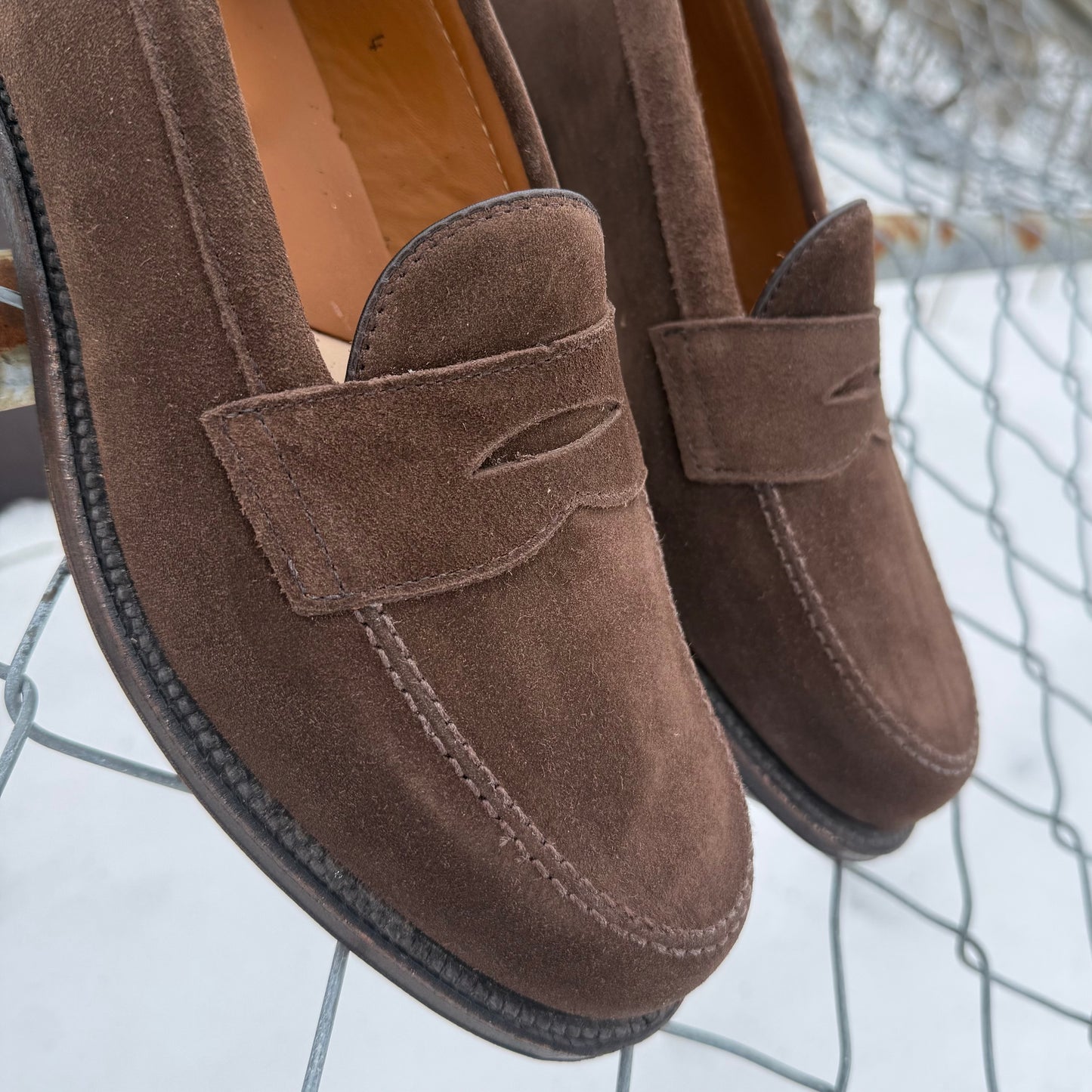 Alden Madison’s 6175F Penny Loafer in Humus Suede sits on a wire fence amid snowy scenery, showcasing Van Last shaping, moccasin stitching, a penny slot detail, and a Flex Oiled Leather Sole for comfort and style.