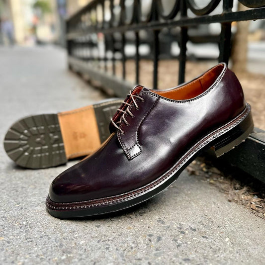 A Close-up of Alden Madisons 990C, a Color 8 shell cordovan leather dress shoe with the Barrie Last design and brown laces, on concrete. Its commando sole is seen as another shoe lies on its side nearby, with a metal fence and blurred urban background in view.