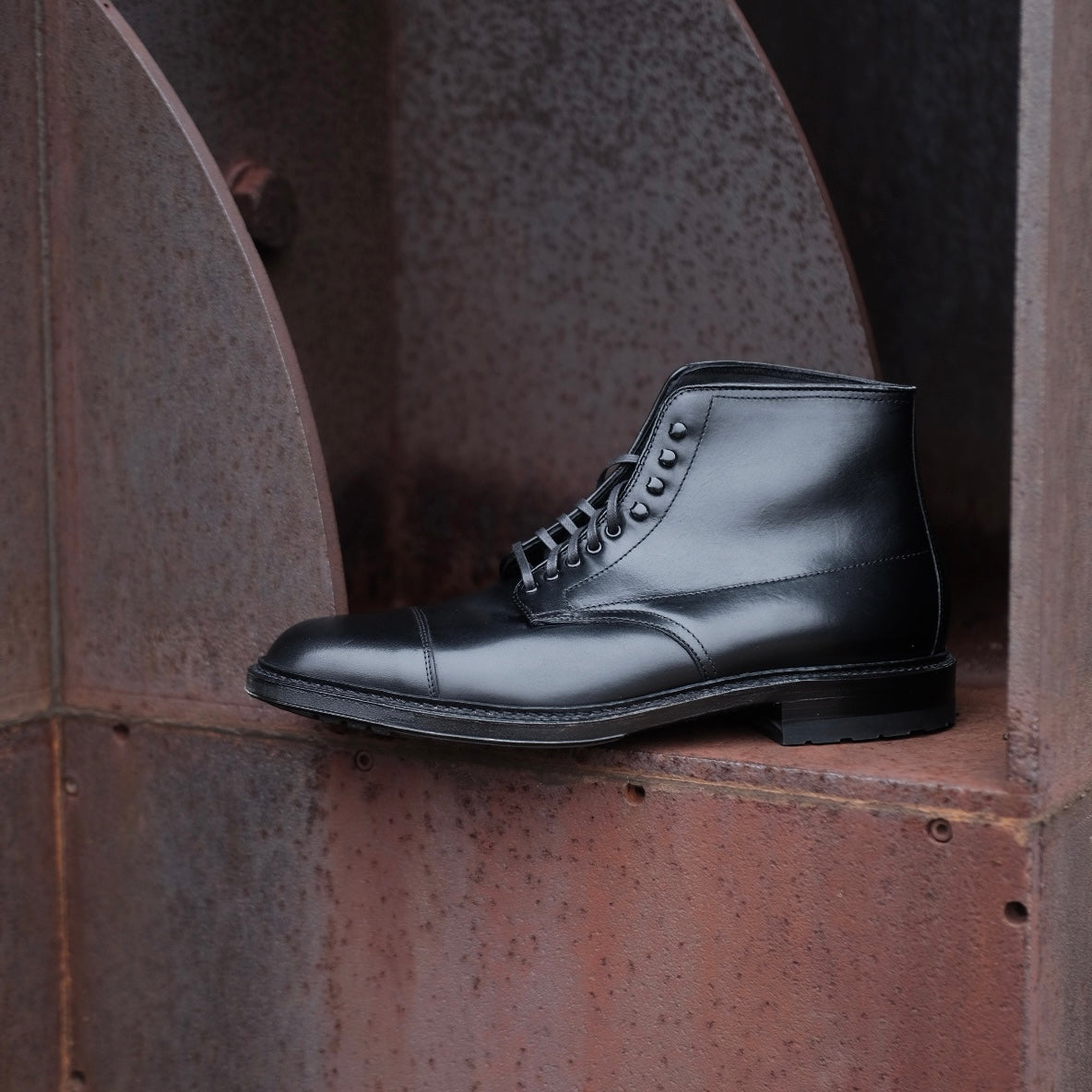 The Alden Madison 40797HC Black Cap Toe Boot with Commando Sole is displayed on a rusty metal surface, set against an industrial backdrop featuring weathered, reddish-brown metal textures.