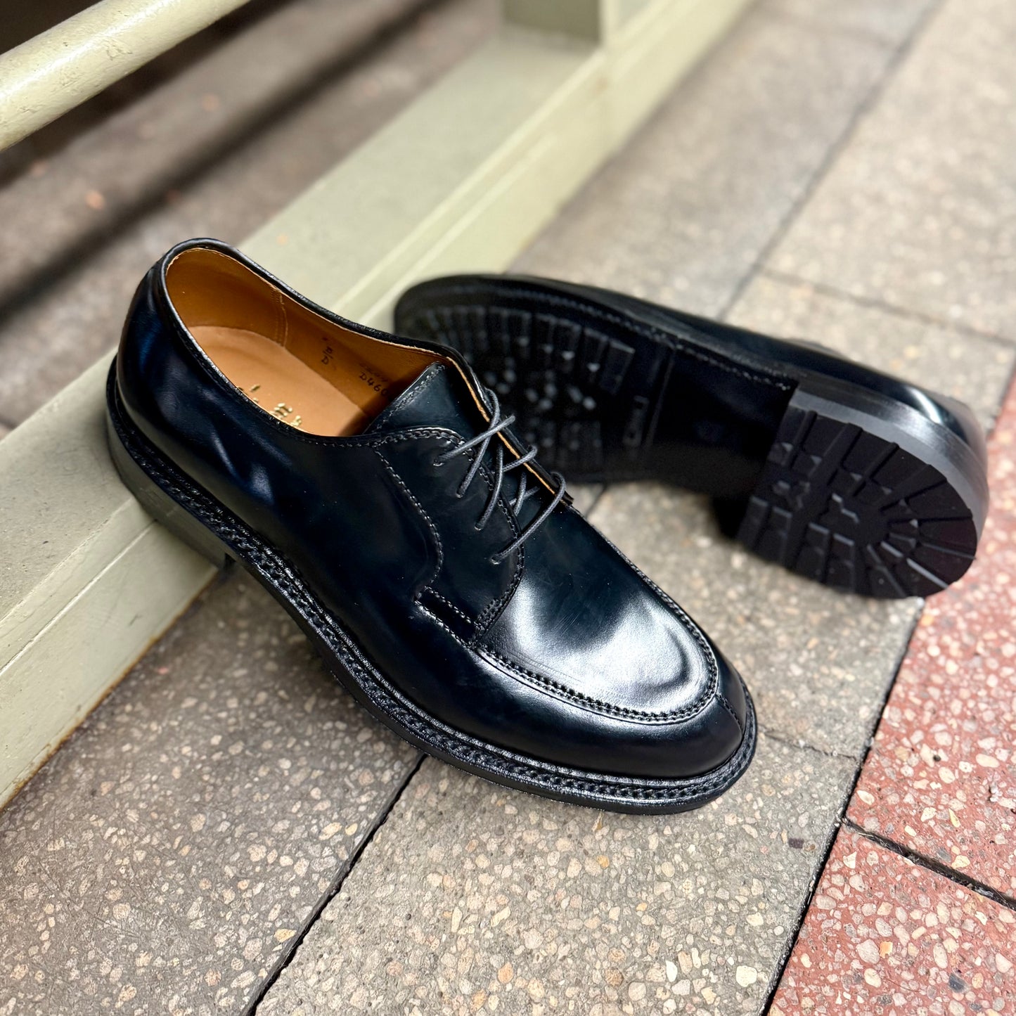 The ALDEN SHOES D4604C - Moc Toe “Borough” Blucher in Black Shell sits on a tiled sidewalk, one upright and one on its side to highlight the shiny finish and detailed tread of the thick sole.