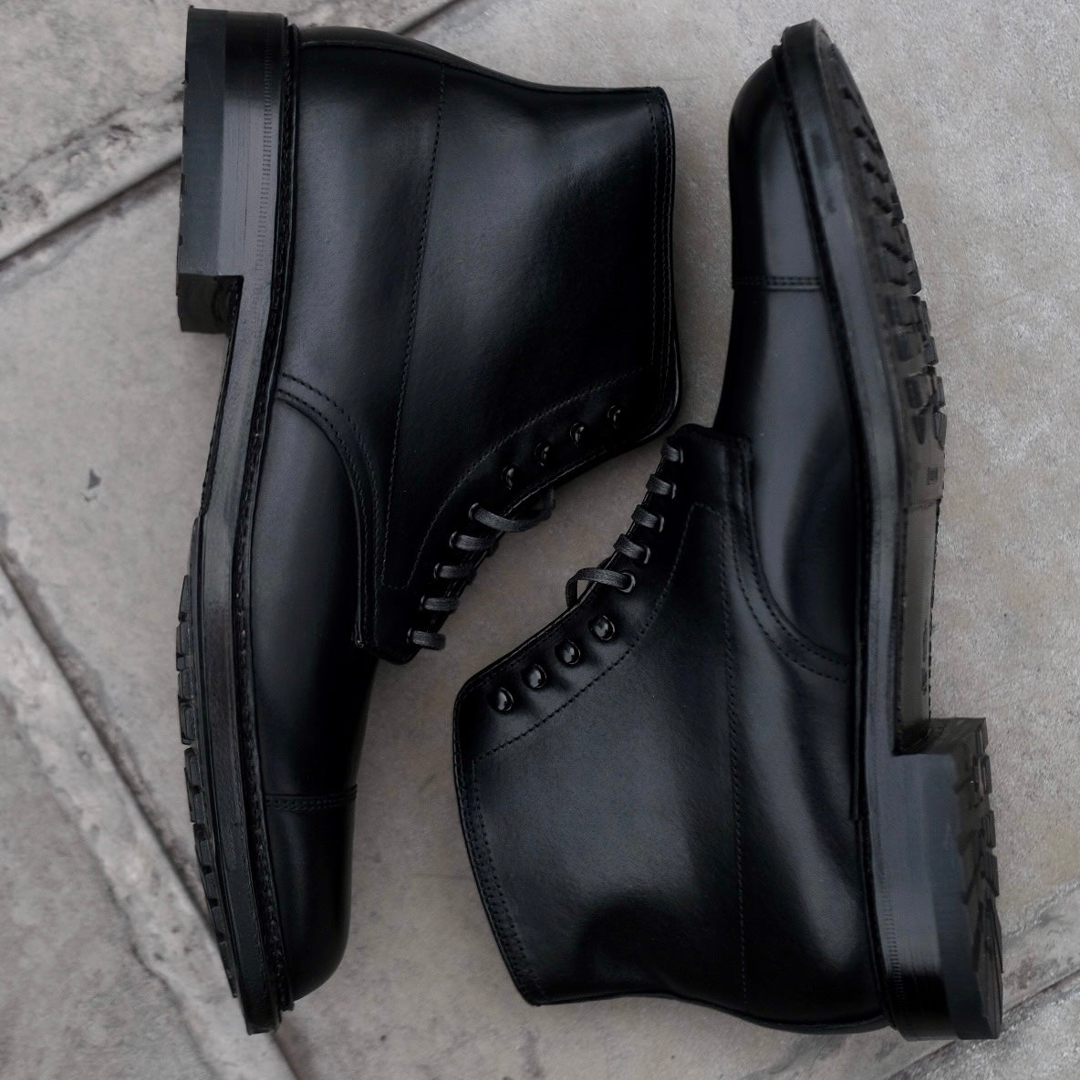 A pair of Alden Madison 40797HC Black Cap Toe Boots with commando soles is displayed side by side on a light-colored, textured stone floor.
