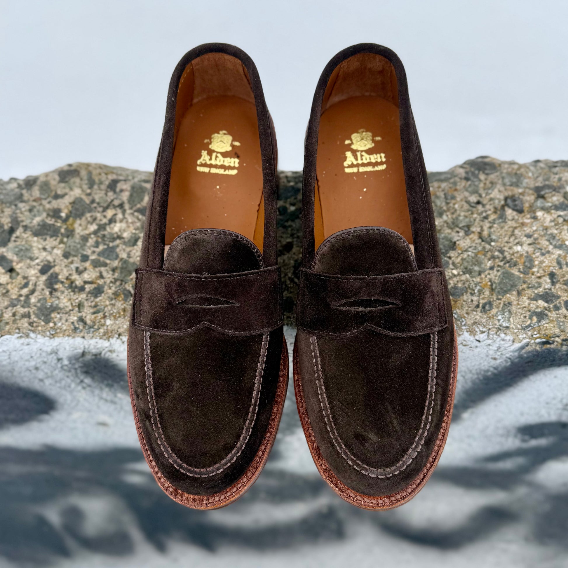 The Alden Madison 6176F Penny Loafer in Dark Chocolate Suede features light brown stitching and flex oiled leather soles, shown side by side on a rough stone surface with a blurred background.