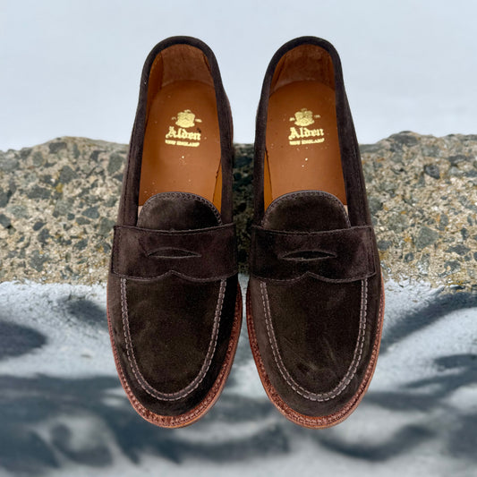 The Alden Madison 6176F Penny Loafer in Dark Chocolate Suede features light brown stitching and flex oiled leather soles, shown side by side on a rough stone surface with a blurred background.