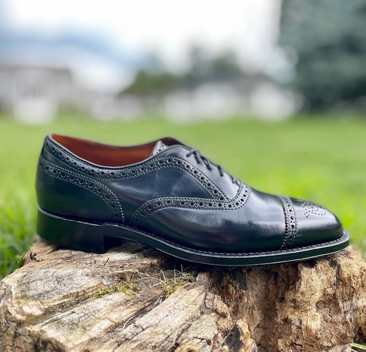 The Alden Madison 9841 - Medallion Cap Toe Bal in Black Shell Cordovan, featuring a Hampton Last and Single Oak Leather Sole, sits outdoors on a tree stump amid green grass and blurred trees.