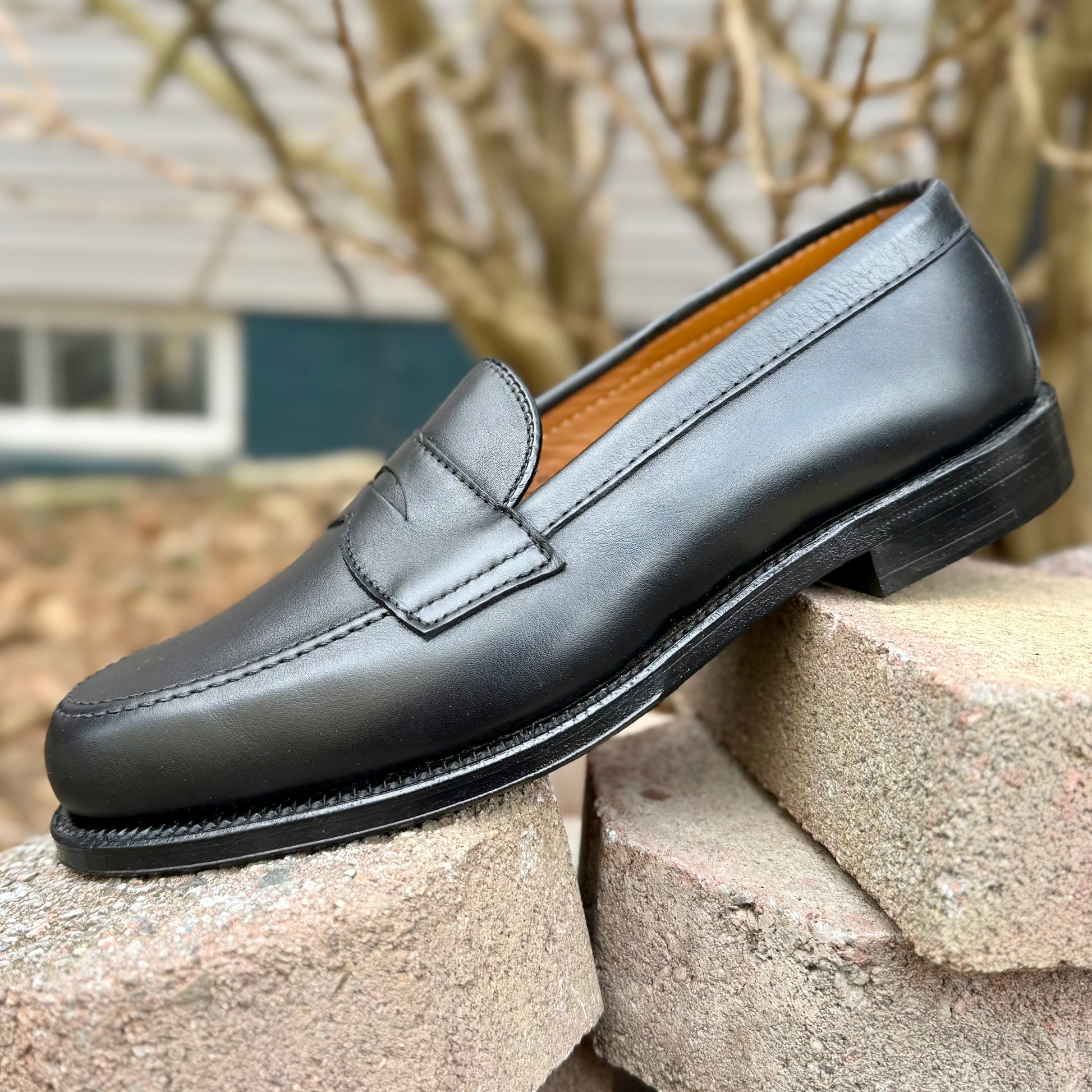 The ALDEN SHOES 9827 - Black Calf Leisure Penny Loafer with a leather sole is displayed outdoors on stacked concrete bricks, set against a blurred house and bare branches in the background.