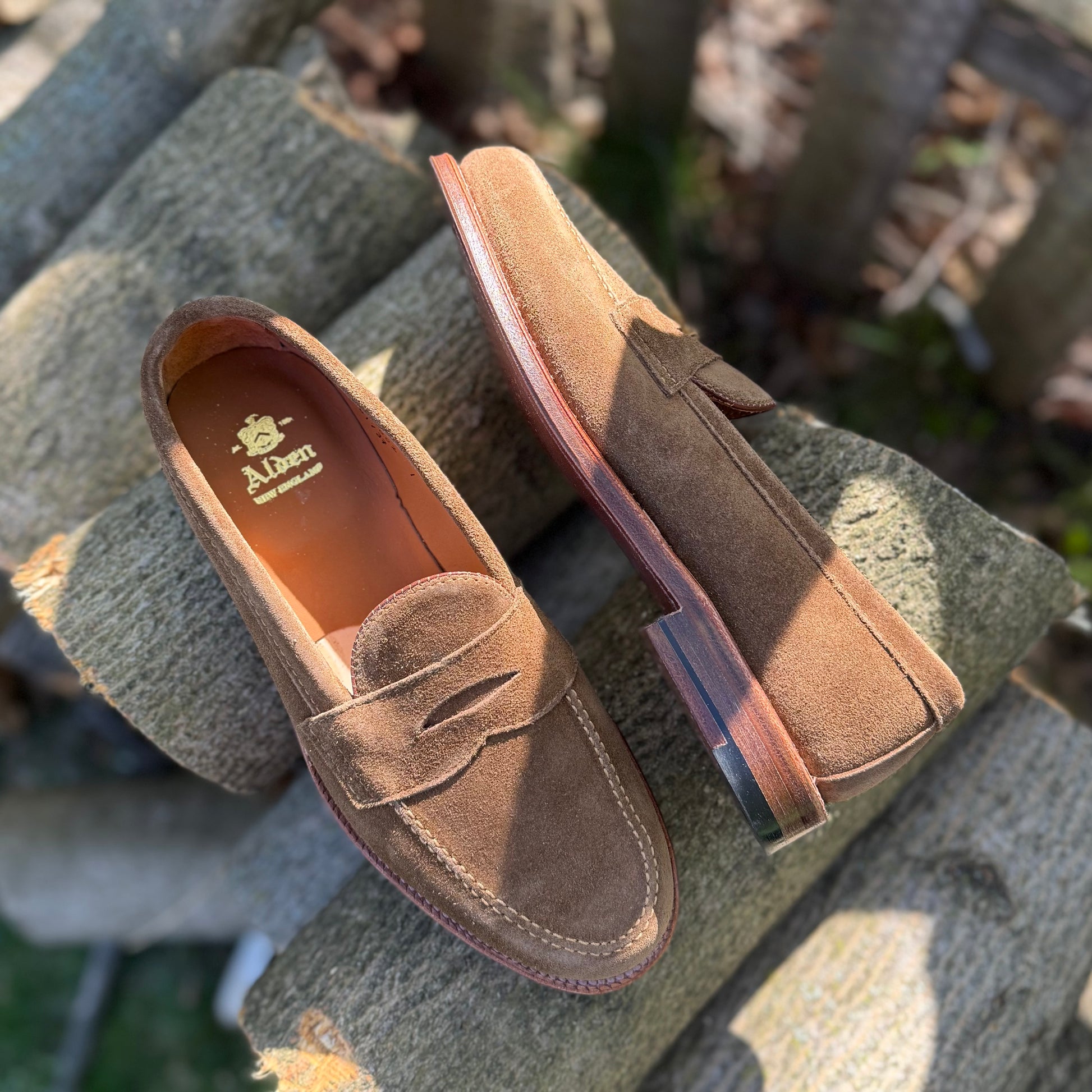 Alden Madison’s 6173F - Penny Loafer in Snuff Suede, crafted on the Van Last with Flex Leather Soles, is stylishly displayed outdoors—one flat and one propped on textured wooden logs.