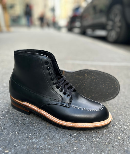 The Alden Madison 401 - Indy Boot in Black Chromexcel, featuring brown Neoprene cork soles, is shown on a city sidewalk—one standing upright, the other on its side to display the textured sole.