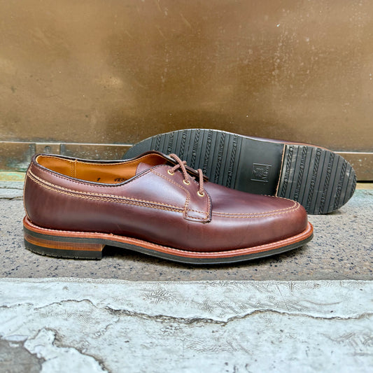 The Alden Madison 734L - Ivy Blucher in Brown Chromexcel is shown, with one shoe upright and the other revealing a black grooved Leisure Rubber Sole, set against a textured sidewalk and metal background.