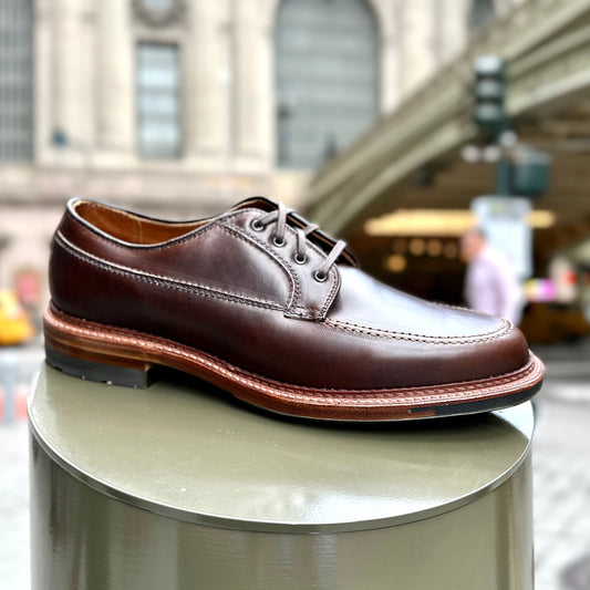 The Alden Madison D3616C - Ranger Mocc in Brown CXL, a polished leather dress shoe with a lace-up design, rugged Commando sole, and contrast stitching, is elegantly presented on a round outdoor pedestal. In the blurred background, an architectural structure and a passerby are visible.