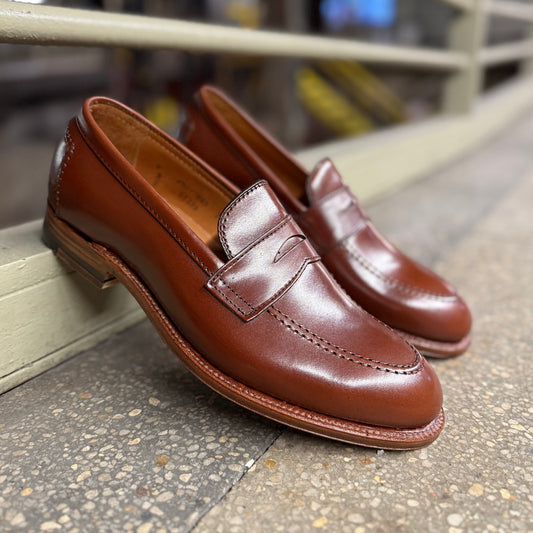 A pair of D1235 - Madison Penny loafers in Madison Brown Calfskin from Alden Madison is showcased on a gray stone surface. The shoes are angled with toes inward, accentuating their polished finish and classic design against a blurred background.