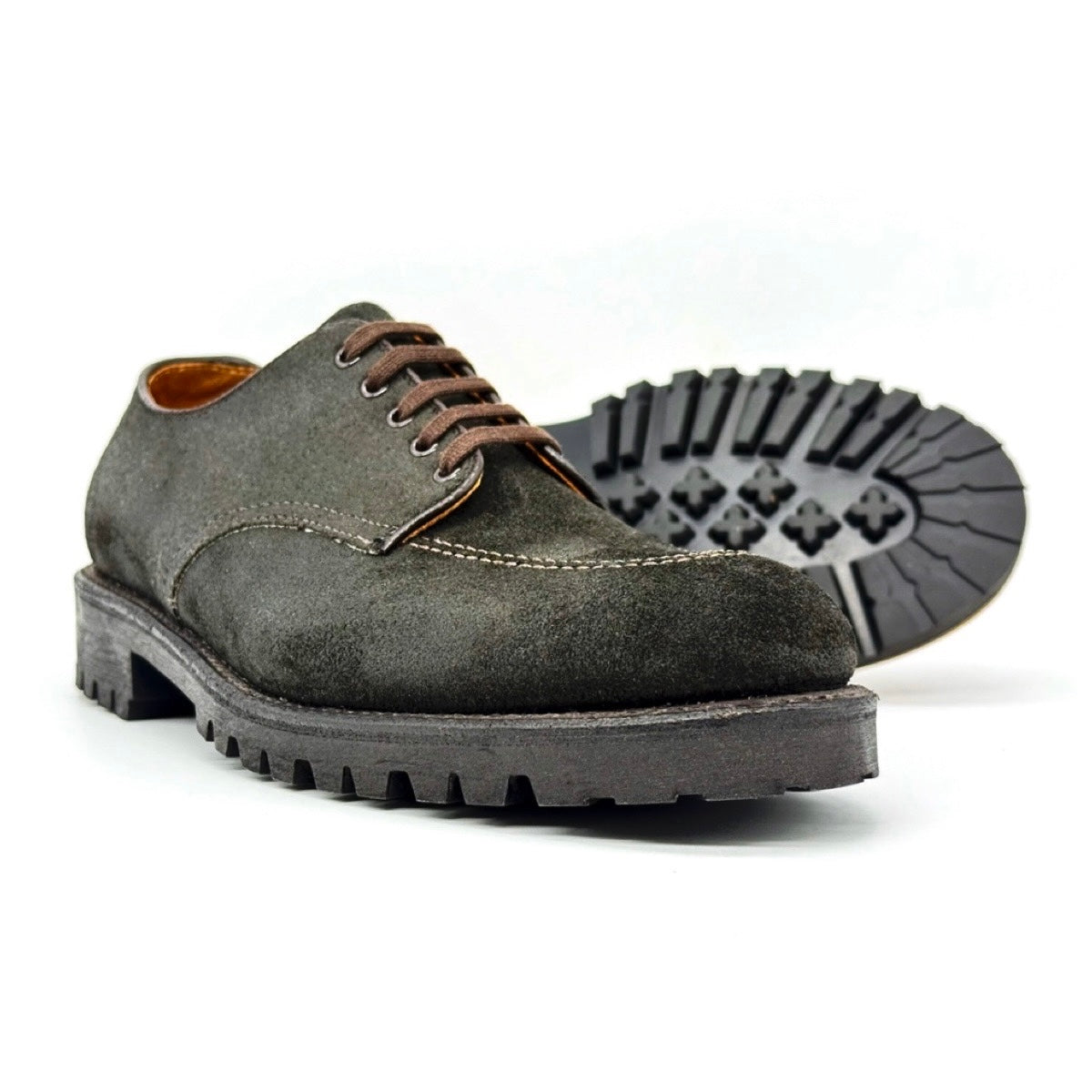 Introducing the ALDEN SHOES 4006 - Earth Reverse Indy Shoe Heavy Lug: a pair of rugged, dark gray suede lace-up shoes featuring brown laces, prominent visible stitching, and thick Heavy Lug Rubber Soles for dependable traction. Expertly crafted on the iconic Trubalance Last, one shoe stands upright while the other is angled to display its distinctive patterned tread—a true homage to the classic Indy Shoe heritage.