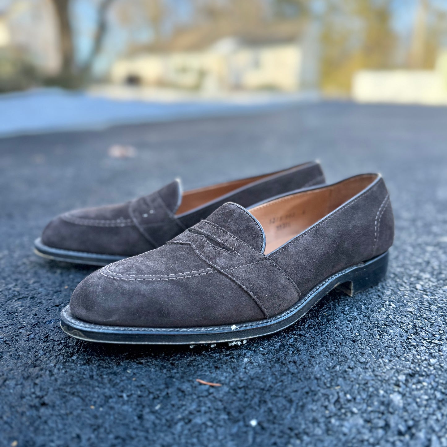The ALDEN SHOES G9201 Dark Brown Suede Full Strap Loafer, featuring detailed stitching, is displayed outdoors on a blacktop surface with trees and houses blurred in the background.