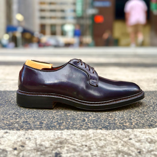 An Alden Madison 990 Plain Toe Blucher in Color 8 Shell Cordovan, made on the Barrie Last, with laces, sits on a crosswalk in a city setting with blurred buildings and a person in the background.