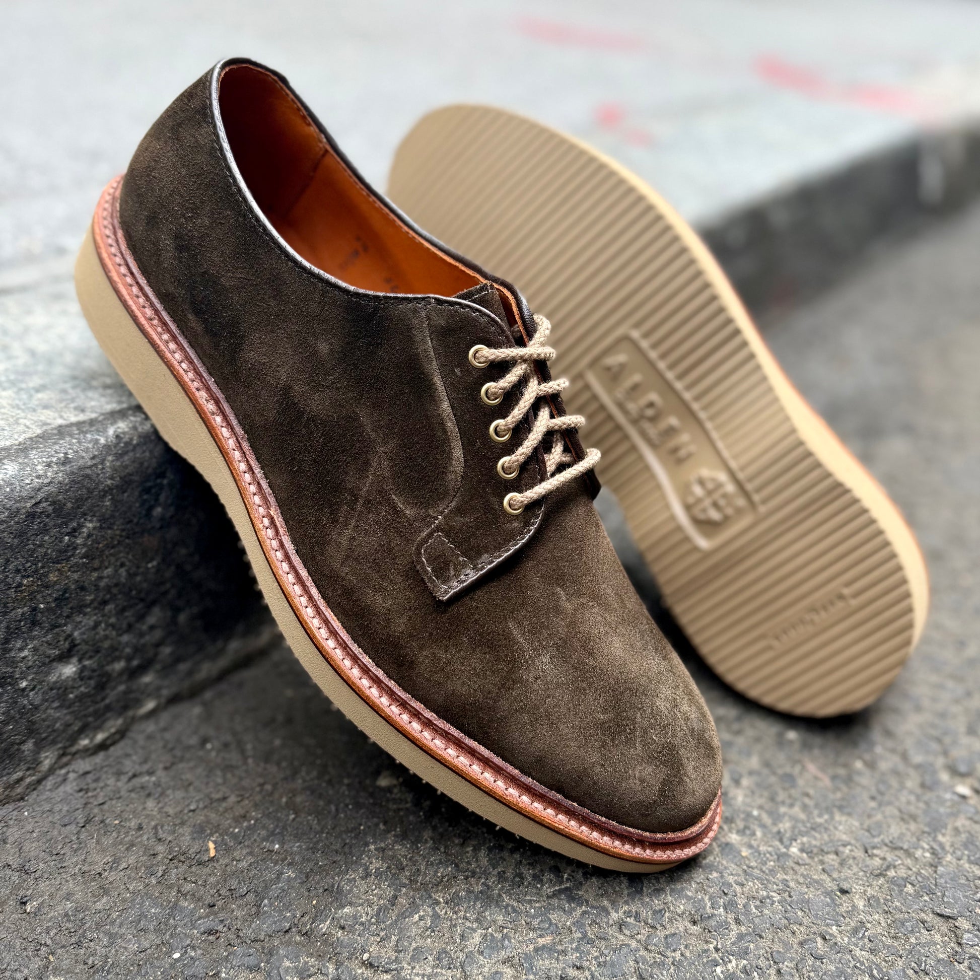 A pair of ALDEN SHOES D5426 - Loden Suede Plain Toe Bluchers in brown suede with light tan rubber soles, shown on rough outdoor pavement—one upright, the other on its side revealing the sole.