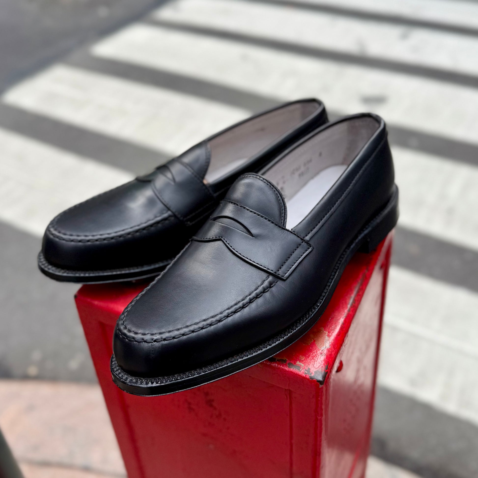 A pair of ALDEN SHOES 9817 - LHS Penny Loafers in Black Trapper Calf are showcased outdoors on a red metal box, with a crosswalk and street softly blurred in the background.