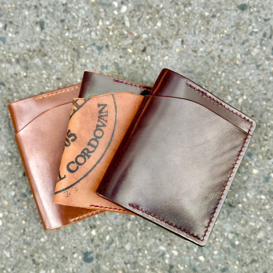 Three Alden Madison Herb Bi-Fold Wallets in Shell Cordovan are displayed on a textured concrete surface. The handcrafted design features brown shades and quality craftsmanship, with one wallet showing a partially visible printed logo.