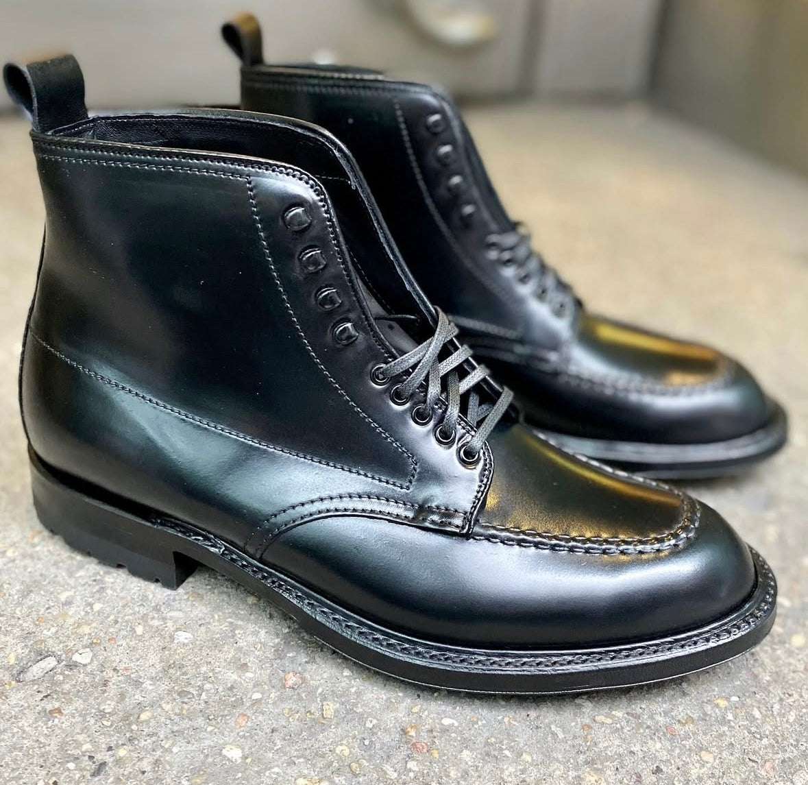 The Alden Madison 40509HC Indy Boot w Commando Black Shell features shiny black leather, a moc toe design, and a sturdy Commando sole, stylishly shown on a textured concrete surface.