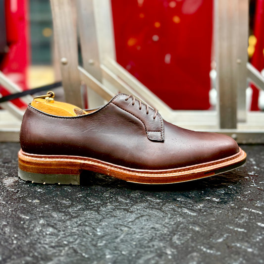 A close-up of the ALDEN SHOES 9506C Plain Toe Blucher w Commando Brown CXL, featuring a thick layered sole, shown on a dark textured surface with a blurred red and metallic background.