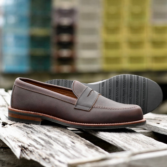 The ALDEN SHOES 5730L Penny Mocc in brown kudu leather with mocc toe stitching and a rubber sole rests on a weathered wooden surface, colorful crates blurred in the background.