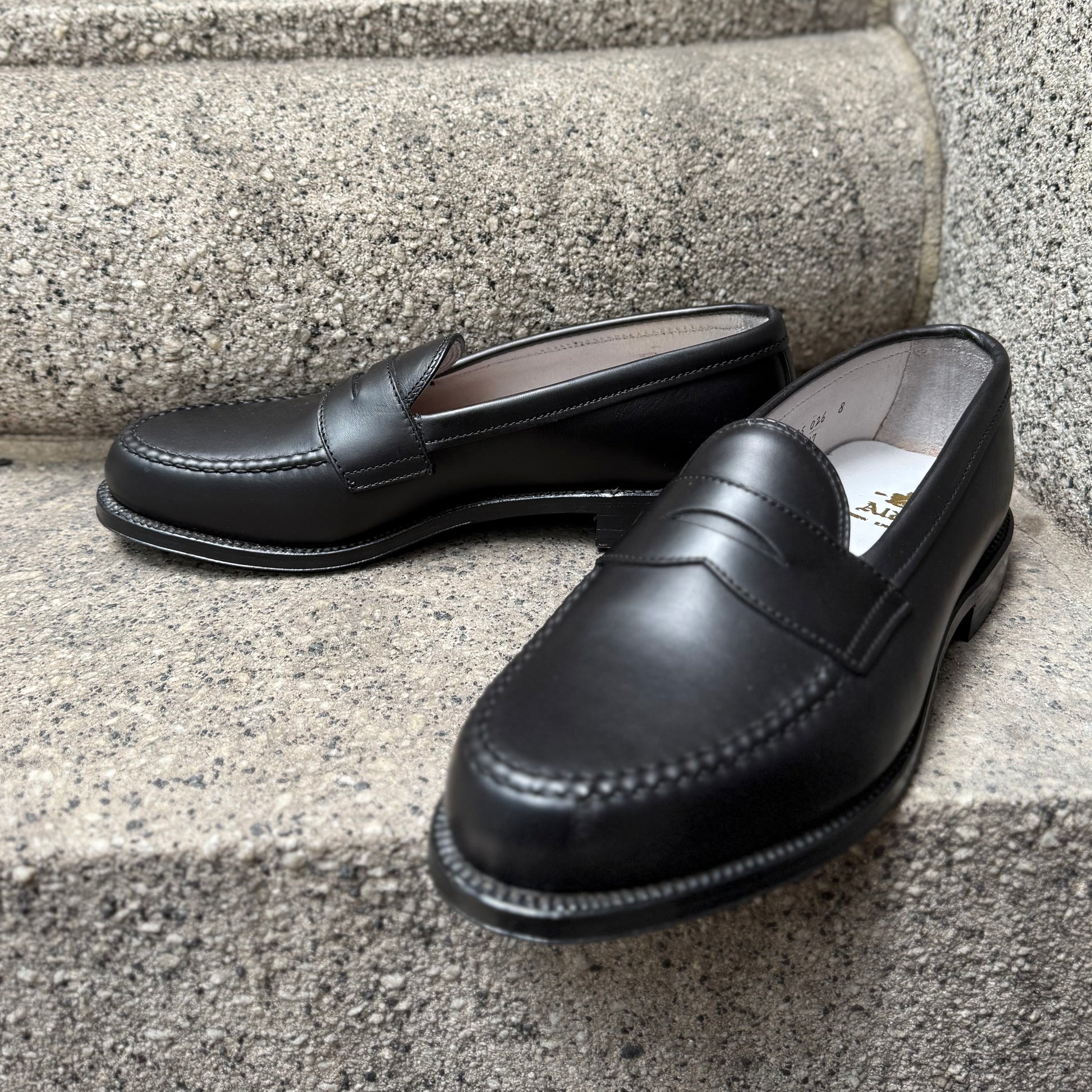 A pair of ALDEN SHOES 9817 - LHS in Black Trapper Calf loafers with hand-sewn stitching is displayed on gray concrete steps.