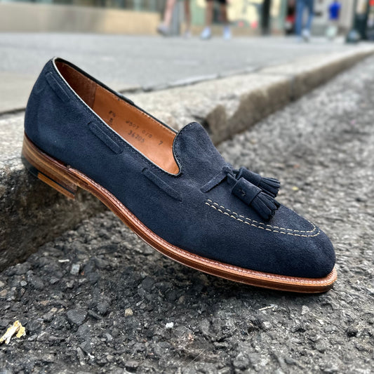 A single Alden Madison 36205 Tassel Loafer in navy blue suede, featuring a leather sole and detailed stitching, sits on a sidewalk curb. Its tassels and light brown sole contrast against the blurred city street backdrop.