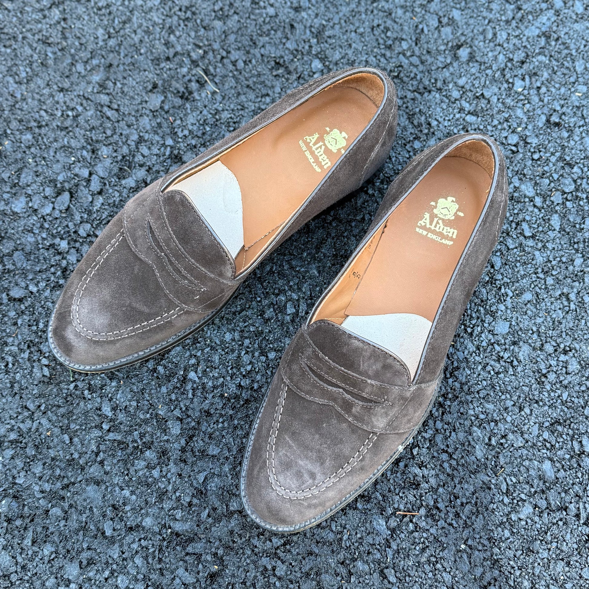A pair of ALDEN SHOES G9201 - Dark Brown Suede Full Strap Loafers with tan leather insoles, featuring the brand's gold logo, placed side by side on textured dark asphalt.